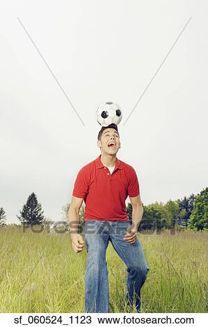 Stock Image - Young man balancing soccer ball on head outdoors. Fotosearch