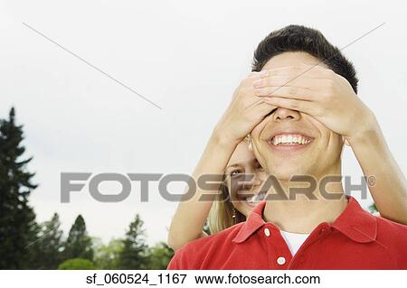 Stock Photo - Young woman covering young man's eyes outdoors. Fotosearch