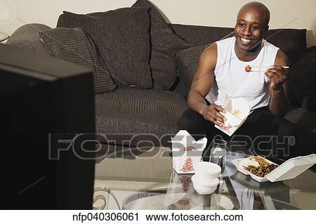 Stock Image - African man on sofa eating take out food. Fotosearch
