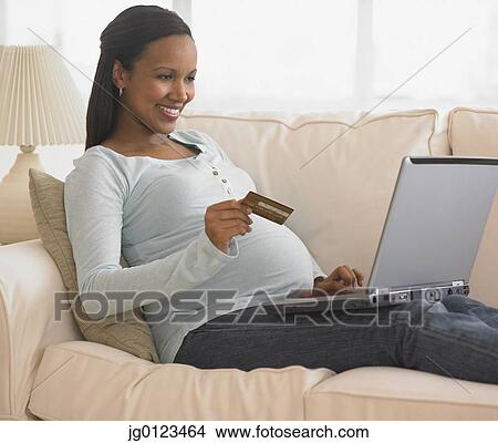 Picture - Pregnant African woman shopping online on sofa. Fotosearch