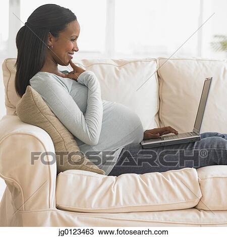 Stock Image - Pregnant African woman typing on laptop on sofa. Fotosearch