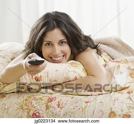 Picture - Hispanic woman laying on bed pointing remote control. Fotosearch