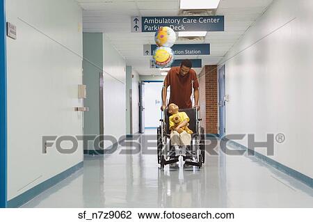 Stock Image - African father pushing son in wheelchair down hospital corridor. Fotosearch