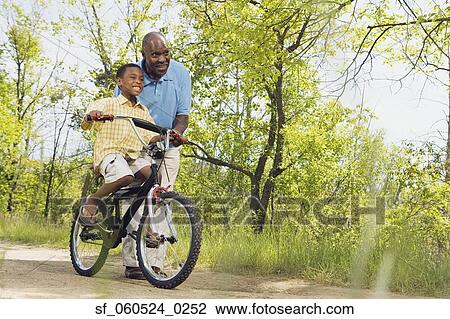 Stock Image - African grandfather teaching grandson to ride a bicycle in park. Fotosearch