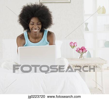Stock Image - African woman using laptop on bed. Fotosearch