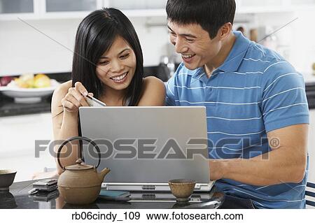 Stock Photo - Asian couple using laptop with credit card in kitchen. Fotosearch