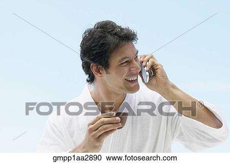Stock Image - Asian man in spa bathrobe drinking tea and using cell phone. Fotosearch