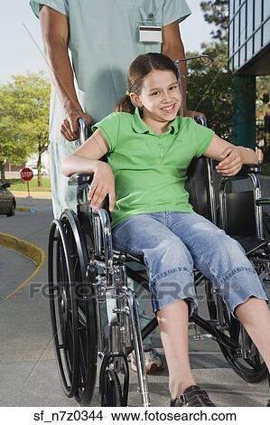 Picture - Girl in wheelchair with nurse on sidewalk next to hospital. Fotosearch