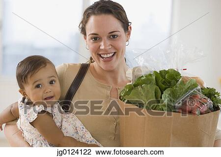 Stock Image - Hispanic mother carrying baby and groceries. Fotosearch