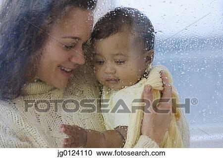 Stock Image - Hispanic mother holding baby behind rainy glass. Fotosearch