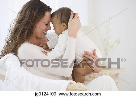 Stock Photo - Hispanic mother sitting in chair hugging baby. Fotosearch