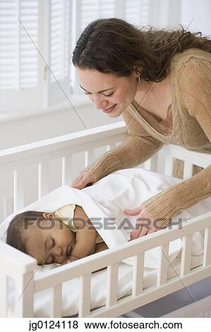 Stock Photo - Hispanic mother tucking sleeping baby in crib. Fotosearch