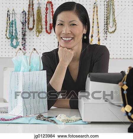 Picture - Middle-aged Asian female boutique owner smiling behind counter. Fotosearch