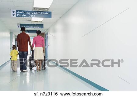 Stock Photograph - Rear view of African family walking down hospital corridor with wheelchair. Fotosearch