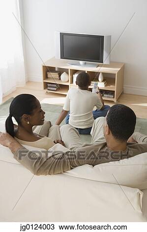 Stock Image - Rear view of African family watching television in livingroom. Fotosearch
