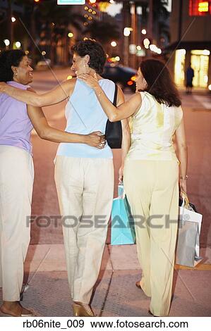 Stock Photo - Rear view of Hispanic women walking on sidewalk with shopping bags. Fotosearch
