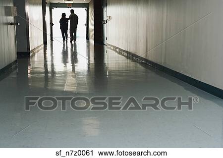 Stock Image - Two people standing at end of corridor in hospital. Fotosearch