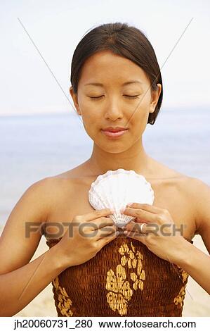 Stock Image - Asian woman holding seashell against chest. Fotosearch