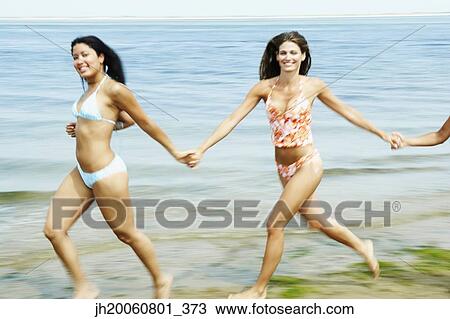 Stock Image - Women holding hands and running at beach. Fotosearch