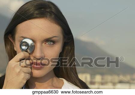Stock Image - Young woman holding stethoscope over eye. Fotosearch