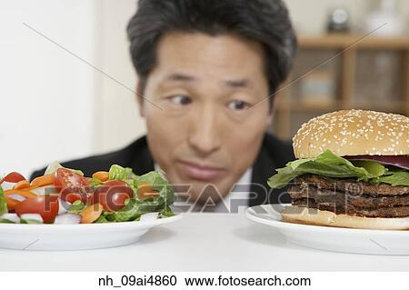 Stock Image - Asian man looking at salad and hamburger. Fotosearch