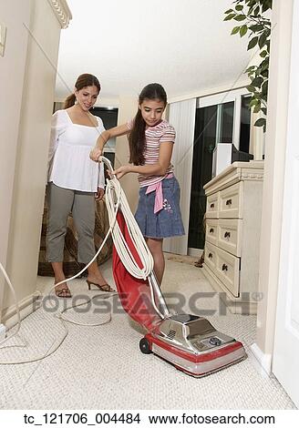 Picture - Hispanic mother watching daughter vacuum. Fotosearch