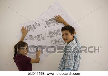 Stock Photo - Asian couple holding blueprints on wall. Fotosearch