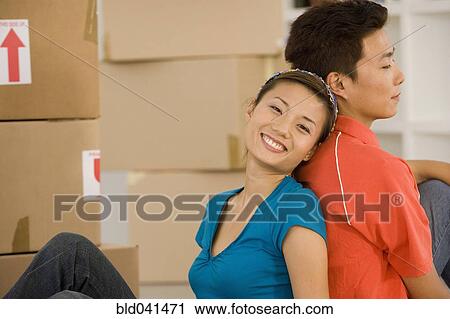 Stock Image - Asian couple sitting back to back next to moving boxes. Fotosearch