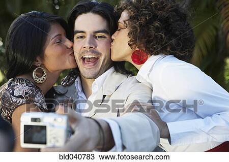 Stock Image - Man taking photograph of two women kissing him. Fotosearch