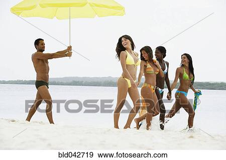 Stock Photo - South American man holding umbrella for women at beach. Fotosearch