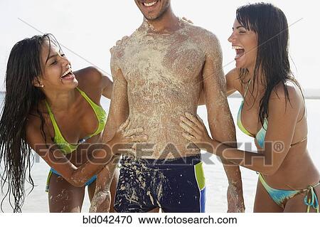 Two women rubbing sand on man at beach View Large Photo Image Stock Image - Two women rubbing sand on man at beach. Fotosearch