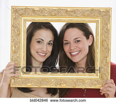 Stock Photograph - Two young women holding picture frame around faces. Fotosearch