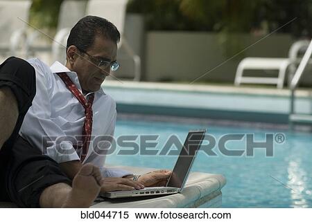 Stock Photography - Hispanic businessman using laptop next to swimming pool. Fotosearch