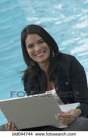 Picture - Hispanic woman with laptop next to swimming pool. Fotosearch