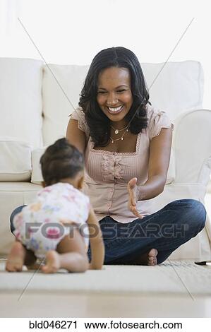 Stock Image - African American mother watching baby crawl. Fotosearch