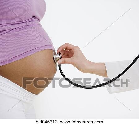 Stock Image - Doctor holding stethoscope on pregnant woman’s belly. Fotosearch