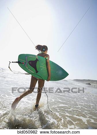 Stock Image - Hispanic girl running with surfboard. Fotosearch