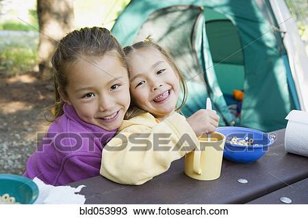 Stock Image - Asian sisters eating at campsite. Fotosearch