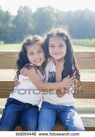 Stock Photograph - Hispanic sisters hugging on bench. Fotosearch