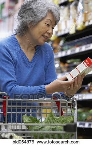 Stock Photograph - Senior Asian woman reading label at grocery store. Fotosearch