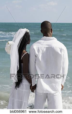 Stock Image - African bride and groom holding hands at beach. Fotosearch