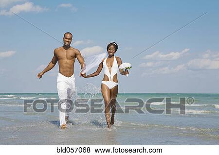 Stock Photo - African bride and groom running in ocean surf. Fotosearch