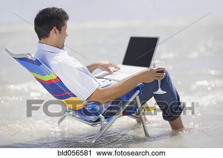 Stock Image - Hispanic man in beach chair with laptop . Fotosearch