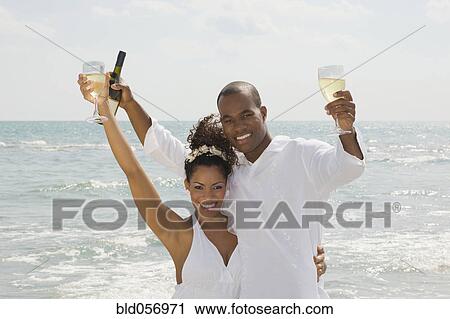 Stock Image - Multi-ethnic couple holding wine at beach. Fotosearch