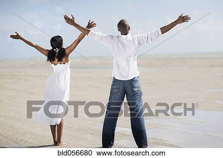 Stock Image - Multi-ethnic couple with arms raised at beach. Fotosearch