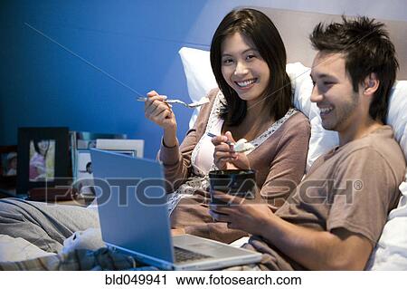 Stock Image - Asian couple eating ice cream in bed. Fotosearch