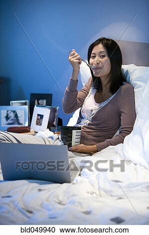 Stock Image - Asian woman eating ice cream in bed. Fotosearch