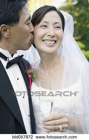 Stock Image - Asian groom kissing bride’s cheek. Fotosearch