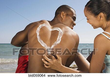Picture - Hispanic woman drawing heart in sunscreen on boyfriend’s back. Fotosearch