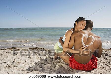 Stock Photograph - Hispanic woman drawing heart in sunscreen on boyfriend’s back. Fotosearch